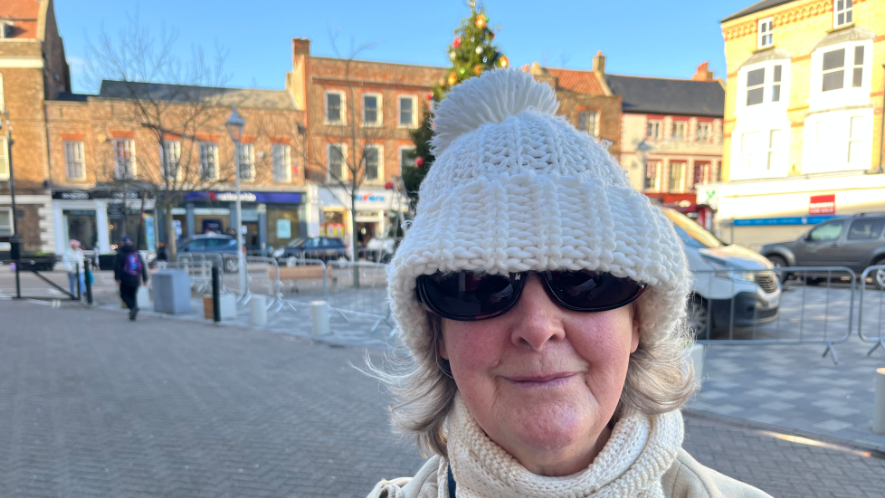 Sue Fletcher is looking at the camera and is wearing a white woolly hat and scarf. She is wearing dark glasses. She is standing in a town square and part of a Christmas tree can be seen behind her.