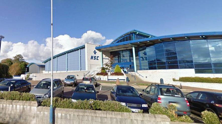 The exterior of the NSC, which is a large glass fronted building with a curved blue roof and a lettering that reads NSC a white wall next to the entrance. There are cars parked in front of the building.