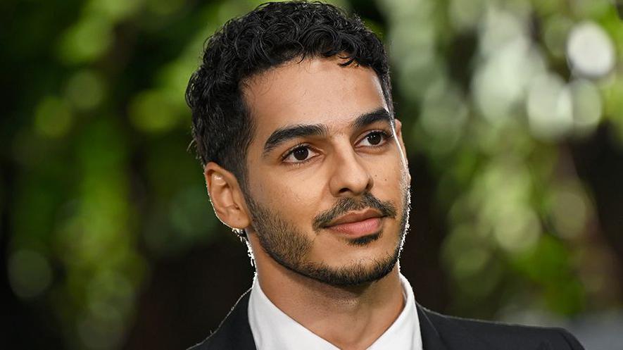 Ishaan Khatter, a male actor at a premiere looking into the distance, wearing a white shirt and black blazer. The background is of green trees blurred and out of focus.