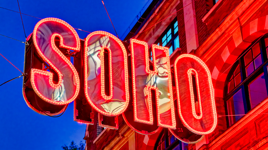 Large red Soho sign with the outline of the letters bordered by small light bulbs. It is a sign attached to a building and it is evening, with dark blue sky in the background.