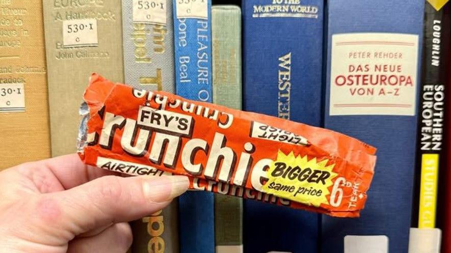 An old chocolate bar wrapper is being held in front of some library books in a woman's hand. The wrapper is orange and has the words "Fry's Crunchie" on it, and the price, which was 6d at the time.