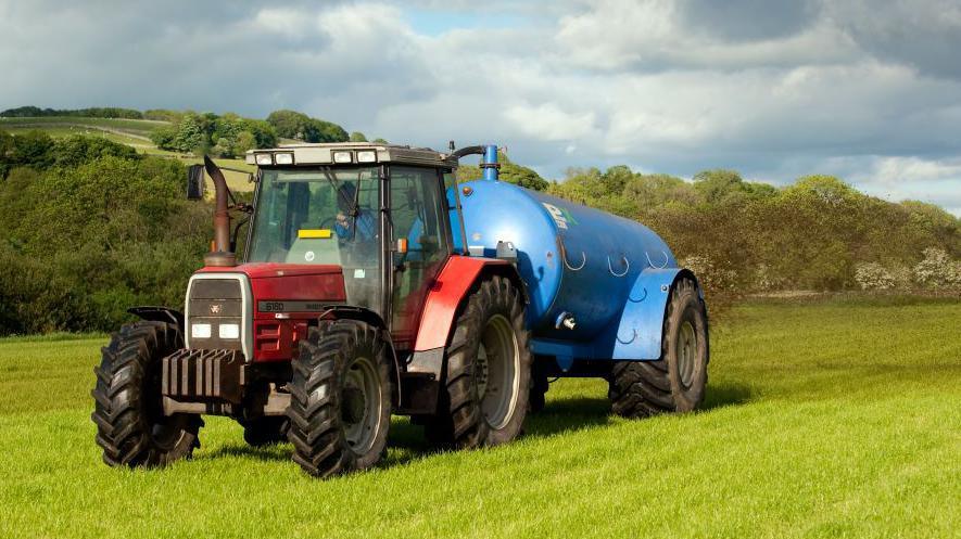 A red tractor in a field with a blue slurry tanker with slurry coming out the back of it. There are blue skies with a few clouds. 