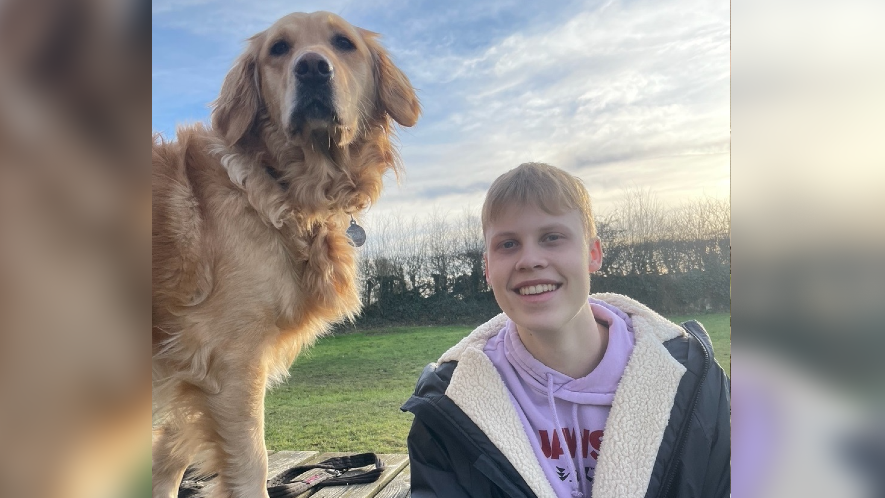 Seb sitting outdoors at a picnic table smiling at the camera, wearing a coat and hoodie. His dog - a golden-Labrador type - is next to him.