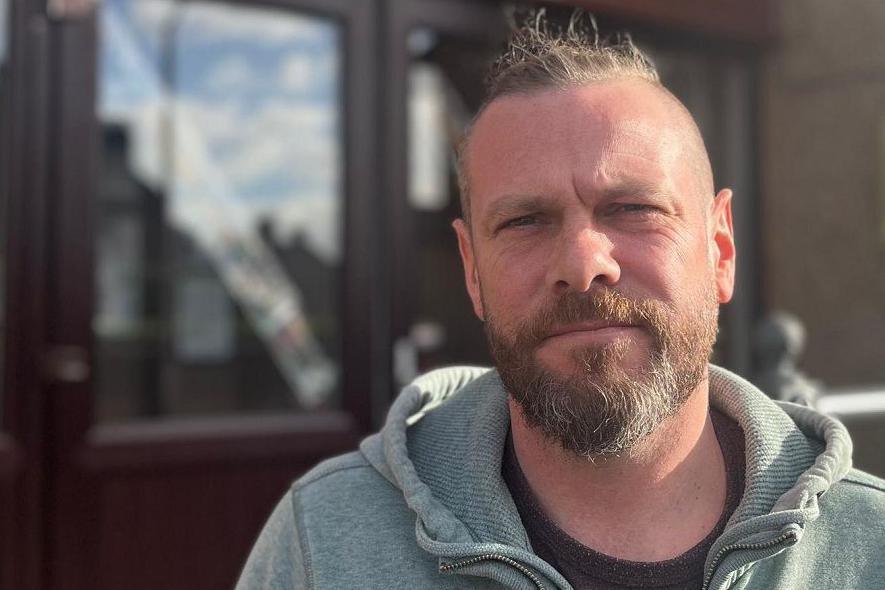 Dave Coulter, who has a beard, is wearing a grey hooded top. There is a door with a glass panel behind him. 