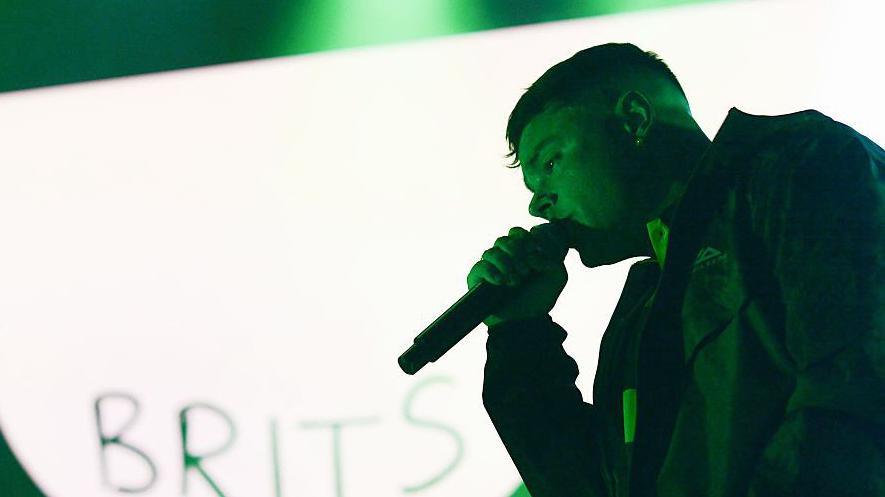 Móglaí Bap of Kneecap performs on stage at the O2 Academy in Glasgow. He is standing in front of a green and white background, with the word Brits written at the bottom corner.