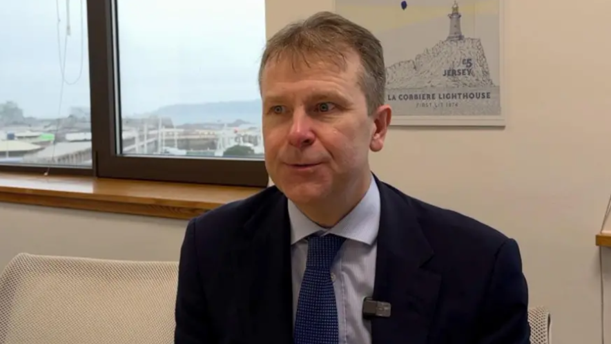 Matt Thomas in a suit with a blue shirt and blue tie looks at the camera, seated in an office. 