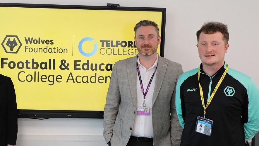 Two men stand to the right of a yellow screen which has the Wolves Foundation and Telford College logos on it. The man on the left is wearing a checked blazer and white shirt and the other a Wolves black and turquoise tracksuit. Both have name badge lanyards on and are smiling at the camera.
