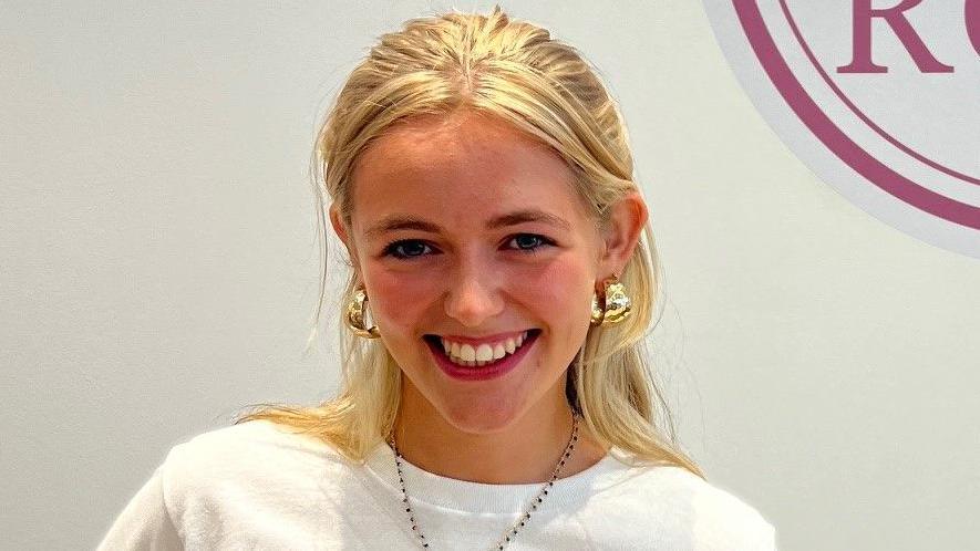A teenage girl with blonde hair standing in front of a white wall. She is smiling and has large gold, hoop earrings in. 