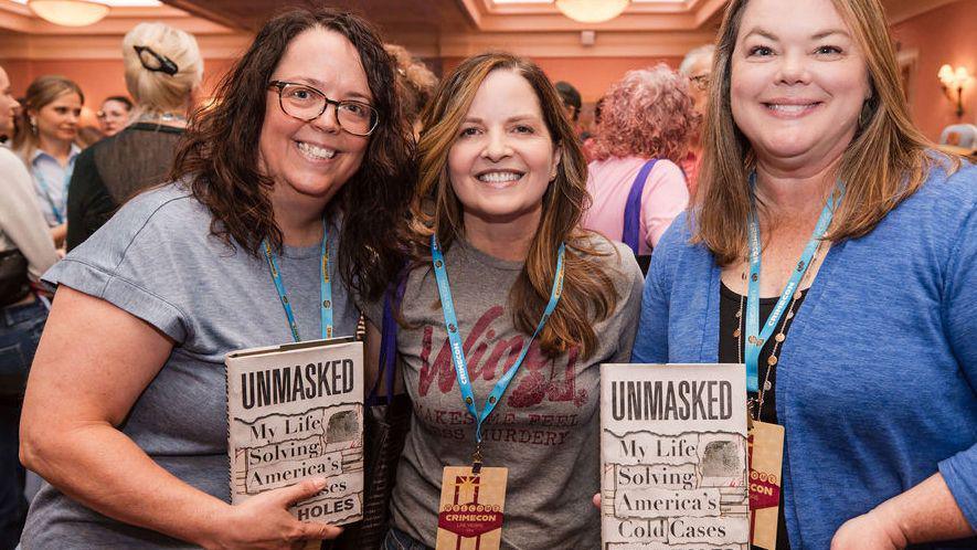 Three female attendees wearing lanyards hold up books titled: "Unmasked - My Life solving America's Cold Cases"