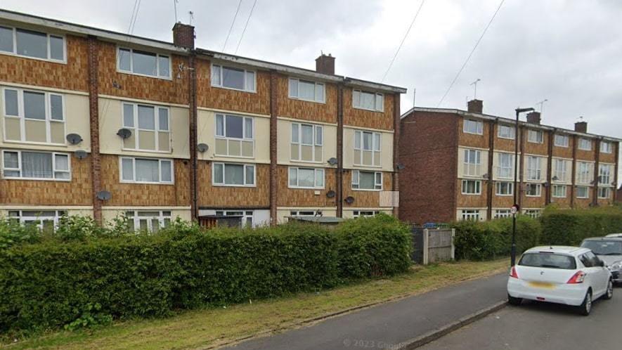 Two four-storey block of flats in Haslam Crescent. The entrance to the homes is obstructed by a row of hedges. Parked cars can be seen lining the road outside. 