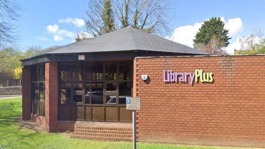 Brick-built hexagonal single-storey library building with many windows. A brick to the right has the words Library Plus in pink and yellow lettering. There are trees and grass around the library.