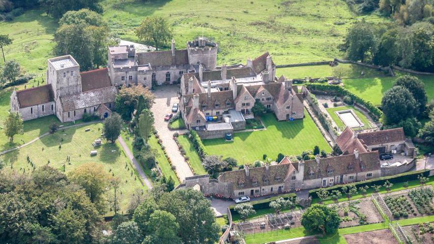 An areal view of Lympne Castle showing the outer grounds and estate. There is green grass and grey buildings. 