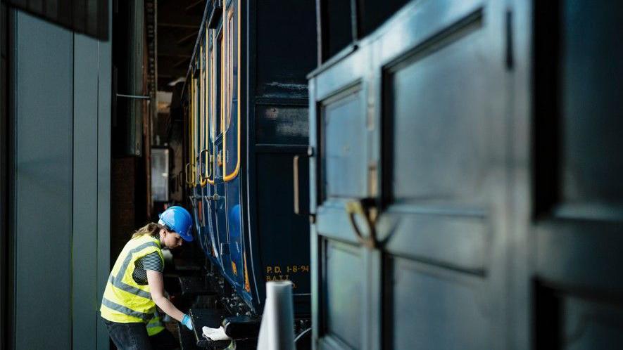 A woman, who is wearing a high-vis jacket with the words Science + Industry Museum on the back, is cleaning a carriage within the museum. 