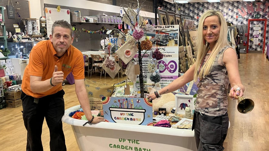 Dave Poulton wearing an orange T-shirt and black trousers standing inside the unity store, beside an upcycled bath tub - with his partner Kez Hayes-Palmer, standing on the other side of the bath tub - wearing a military print sleeveless shirt and brown trousers.