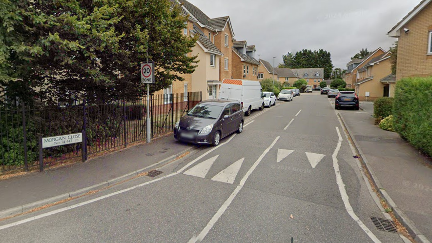 Morgan Close in Luton. The road is a residential area with houses on either side and is a 20mph zone which is signposted. Cars and vans are parked on either side of the pavement of the road.