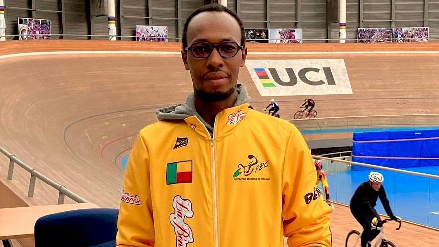 Adrien Niyonshuti, wearing a yellow training jacket with the Benin flag on the right breast, faces the camera as a velodrome with two cyclists on it is seen in the background