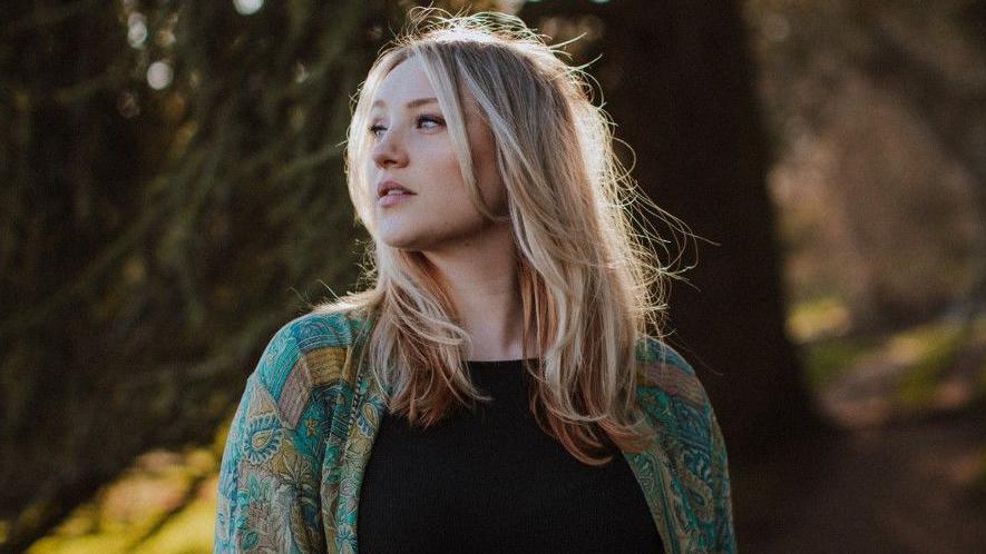 Dolly Mavies in a forest. She wears a black top and a green patterned cardigan. Her hair is lightly blowing in the breeze.