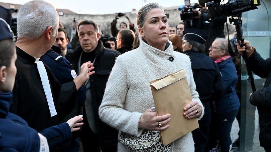 Caroline Darian, wearing a cream coat, walks through a crowd, holding a brown envelope. She is followed by her brothers and there are cameras to her left.