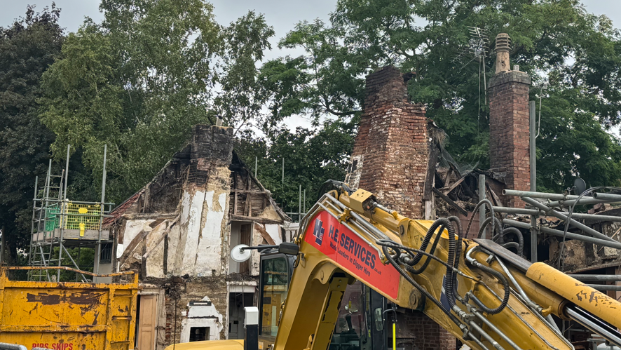 The roof of a cottage in the distance is destroyed. Brick chimneys remains. A yellow skip and part of a yellow crane can also be seen. 