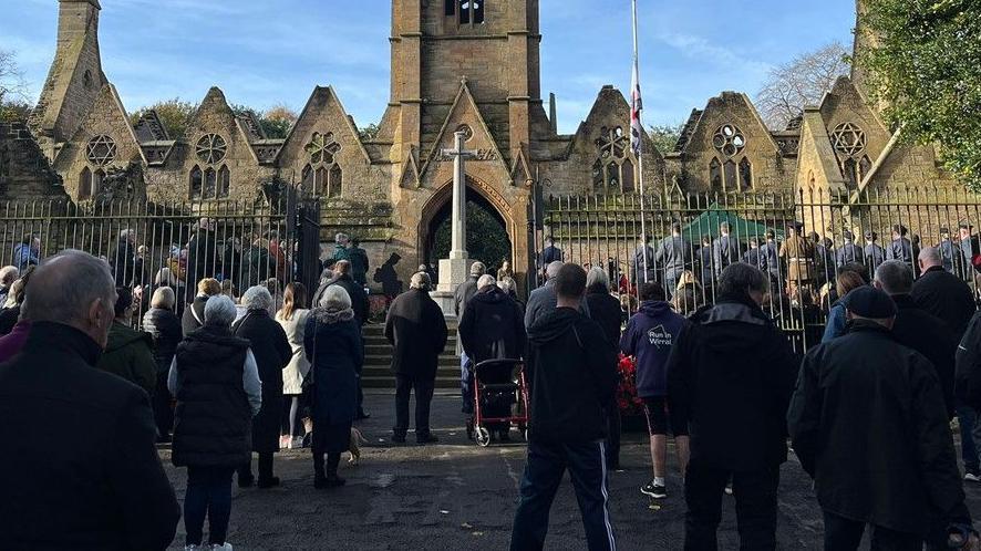 A Remembrance ceremony at Flaybrick Memorial Gardens in Birkenhead, with crowds standing solemnly in front of a church