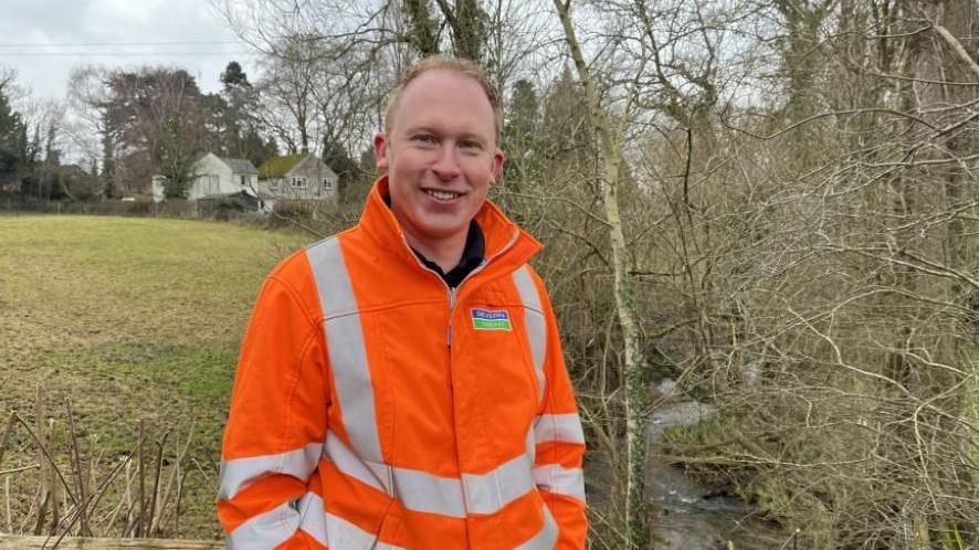 Matt Leedham standing in an orange high vic next to the River Chelt, a tributary of the River Severn. His is stood in a field with trees and homes in the background.