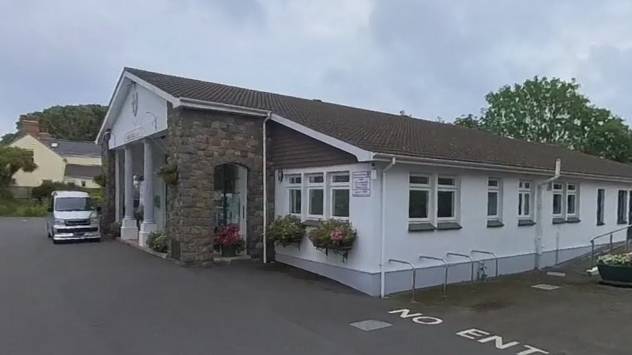 A picture of the front entrance to Vale Douzaine. It is a long white building with a path outside the front and a large silver van parked outside.