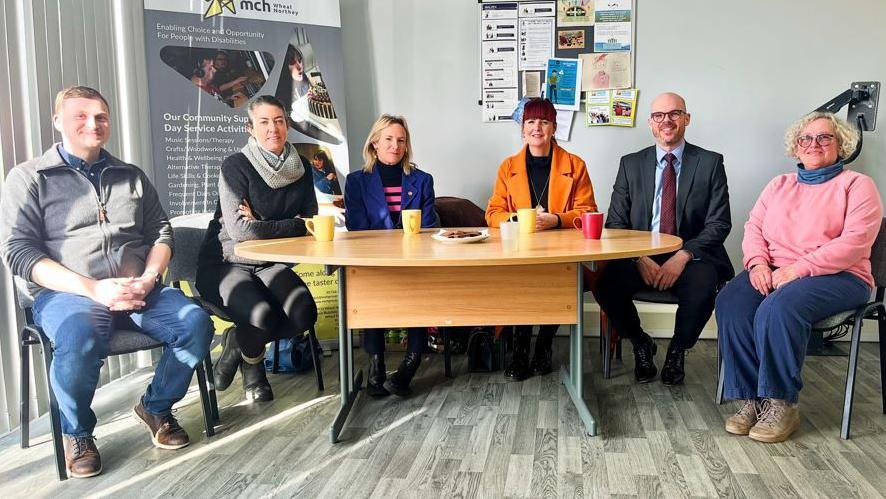 Six people - two men and four women - sat round a table in a room with four mugs on the table along with a plate of biscuits.