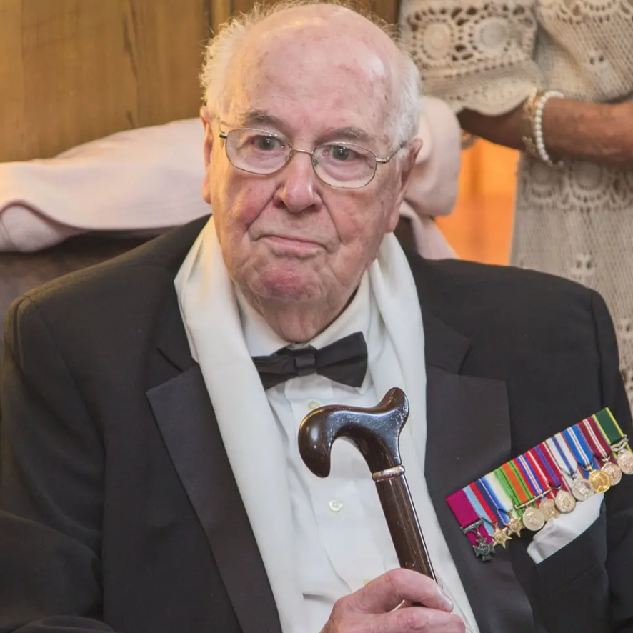 An older John Cruickshank in recent years. He is looking slightly off camera. The top of his head is bald with white hair round the side. He is wearing light-framed glasses and a black suit jacket over a white shirt with a black bow tie. He has a collection of colourful medals on his upper left chest and is holding a walking stick in his right hand.