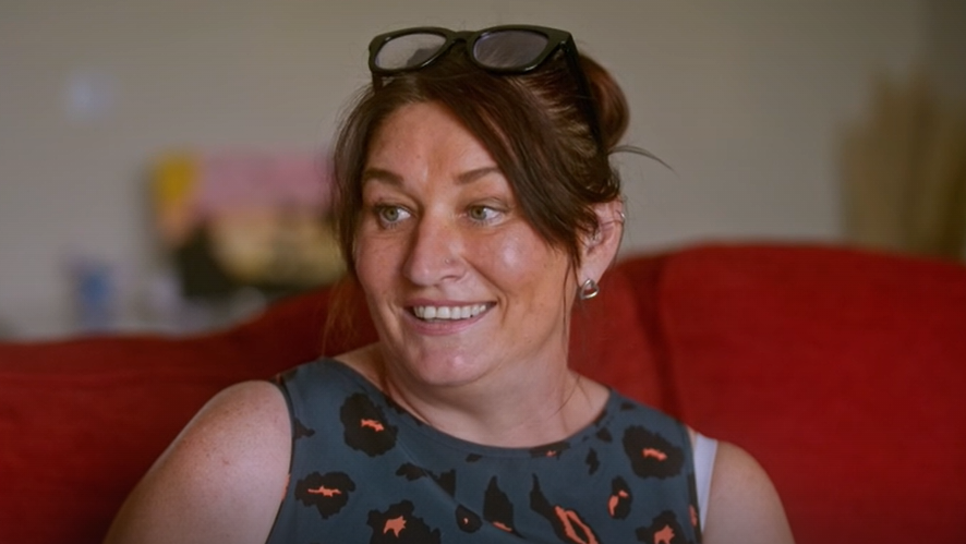 A woman, probably in her 40s, wearing a dark animal print sleeveless top and thick rimmed black glasses perched on top of her head. Her brown hair is tied back and she is wearing gold heart shaped earrings. She is smiling as she sits on a red sofa.