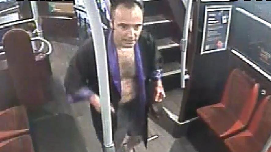 CCTV still of John Baldwin wearing a black kimono with a purple trim. It is open so you can see his chest and he is walking along the lower deck of a bus.