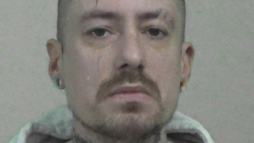Mugshot of Phillips. He has a short beard and shaved head, with tattoos on his neck and large round earrings in the lobes of his ears