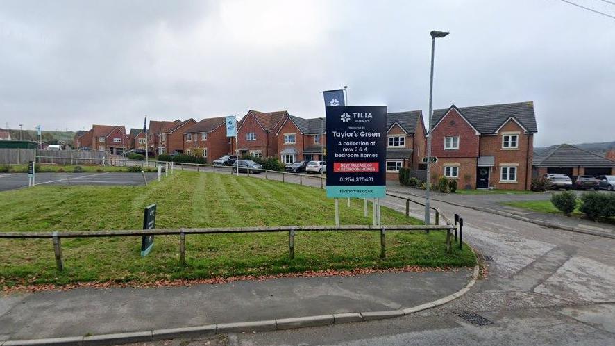 Google streetview of detached red-brick homes on the new estate. They stand alongside a new road, with a grassy area fronting the entrance to the estate and a develops' board advertising the new properties.