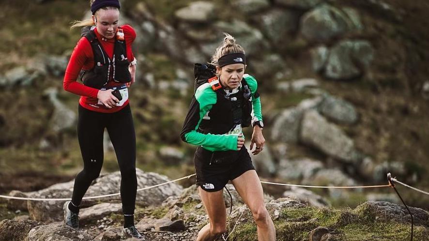 Two women running over rocks with one in black and green gear and another in black and red.