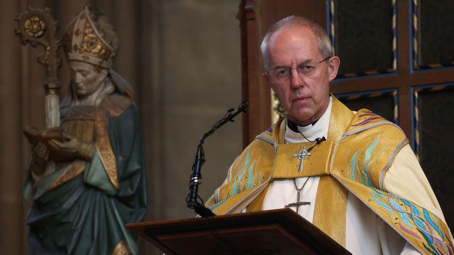 Justin Welby, wearing yellow religious robes, stood at a podium. There is a statue of a figure, also wearing robes, in the background and Welby has a stern expression.