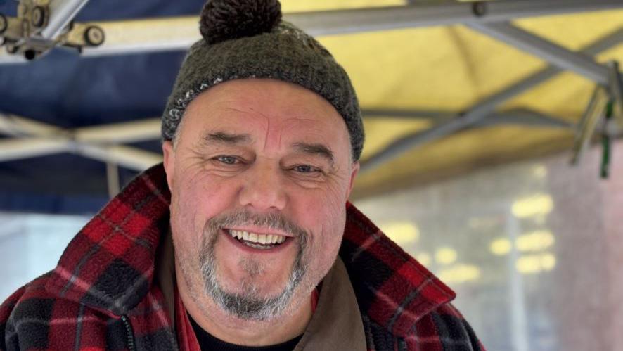 A man wearing grey woolie hat and red and black chequered jacket smiles into the camera.