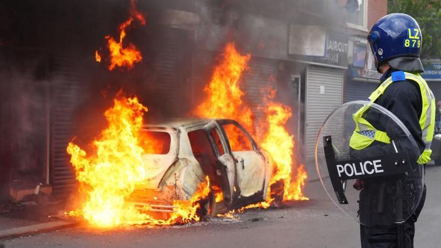 A police officer looks on at a burning car