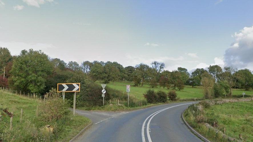 A section of the A523 in Waterhouses, near its junction with Stony Lane - on the left-hand side. The road is seen bending to the right, and there is chevron signage indicating the sharpness of the bend. It is a rural road, with fields and trees surrounding it, bounded by dry stone walls.