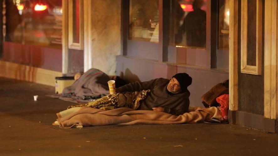 Two people rough sleeping. At the centre of the image is a man lying on a blanket, wearing a hoodie and a black beanie holding out a collection cup. Behind him is another person asleep on the ground. 