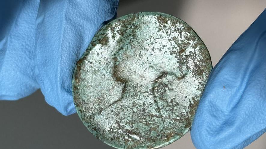 The brass coin has a silver, greenish tinge and is well-worn but the head of the queen is clearly visible. It is being held by a scientist wearing blue plastic gloves. 