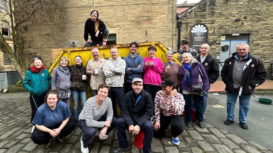 A group of people gathered outdoors on a cobblestone street in front of a large yellow skip placed against a tall brick wall. One person is sitting on top of the skip, while the rest stand or crouch in front of it. The surrounding area features old stone buildings with arched windows and doors, and a leafless tree on the left side.