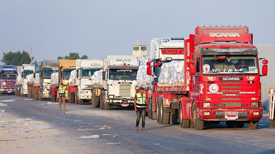 Wide angle image showing a line of eight aid trucks queued at Gaza's Rafah border crossing on Sunday. Two men in hi-vis jackets are stood alongside them