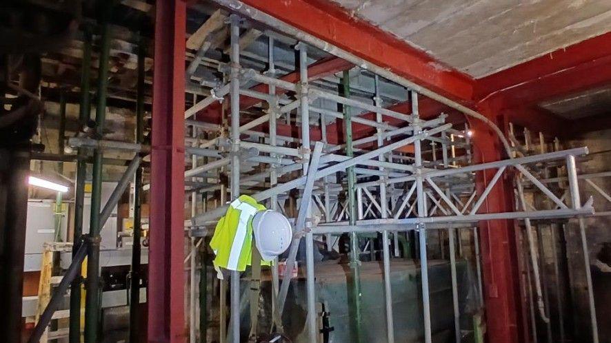 Metal scaffolding poles are supporting red metal girders across the ceiling of a building. There is a yellow high-visibility jacket and a white helmet resting on one of the poles.