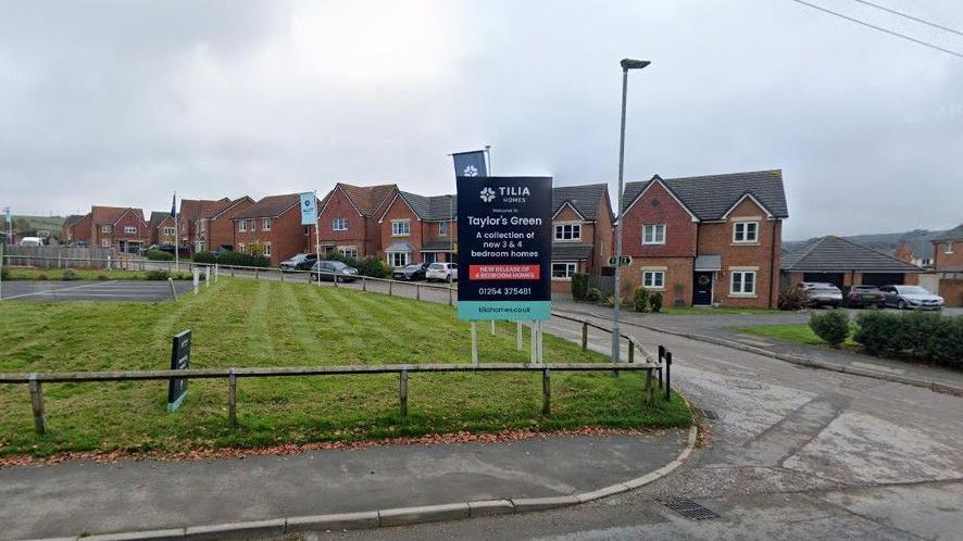 Google streetview of detached redbrick homes on the new estate, lining a road next to a green patch of grass.