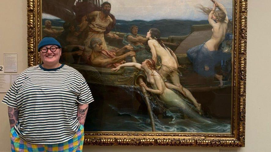 Artist Maddie Morris wearing a black and white striped t-shirt, a blue hat and glasses, with tattoos on their arms. They are standing in front of a large oil painting of Ulysses and the Sirens in an art gallery.