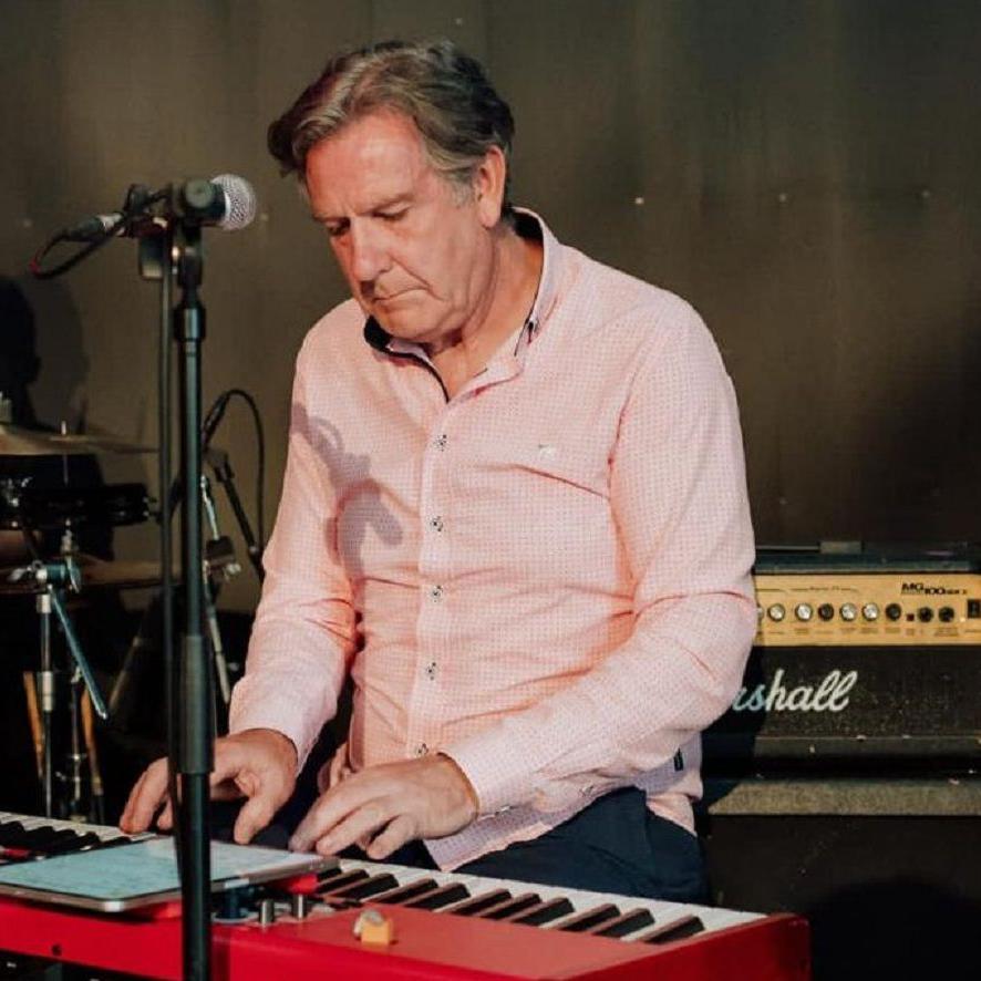Stewart Solley wearing a pink shirt playing a piano. There is a microphone in front of him and an amplifier behind him