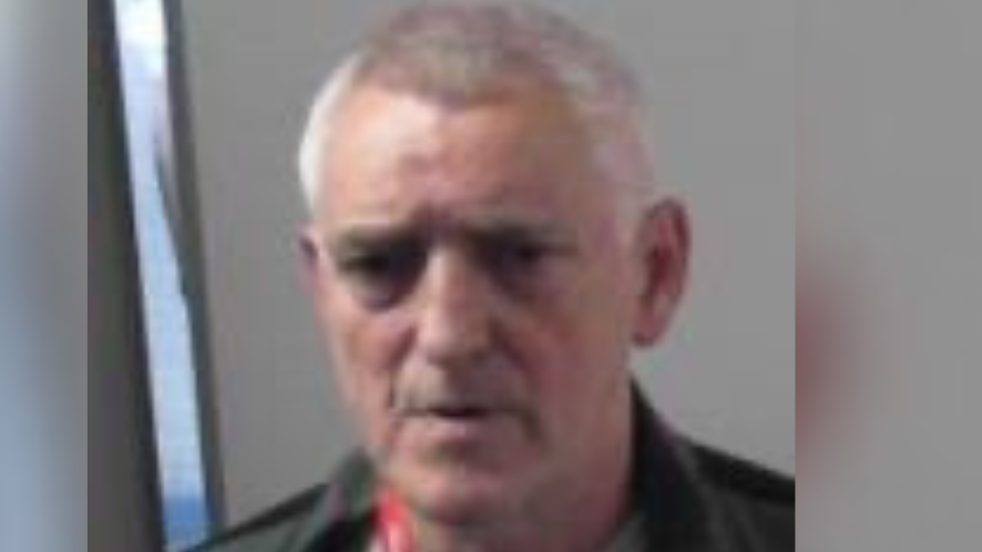 Thomas McCabe - A police mugshot of a man with grey hair, wearing a red lanyard, a green overshirt and white tshirt. He is standing in front of a plain wall.