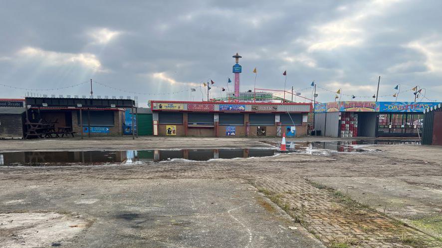 The former Leisure Island Fun Park site. There are puddles of water on the concrete and booths and shacks are now closed with shutters pulled down. 