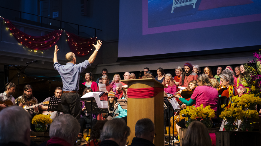 The Bristol Recovery Choir singing at a concert. They are being conducted by a man in a blue shirt and are wearing brightly-coloured tops