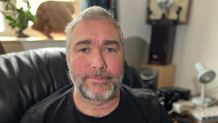 John Goude is looking at the camera and wearing a black T-shirt. He is sitting on a leather sofa in his living room and has greying hair and a beard