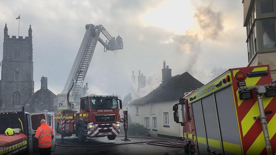 Multiple fire crews are pictured in the village. One fire engine is positioned on support stilts while a ladder extends from the rear of the vehicle so firefighters can squirt water over the thatched-roofed cottages. 